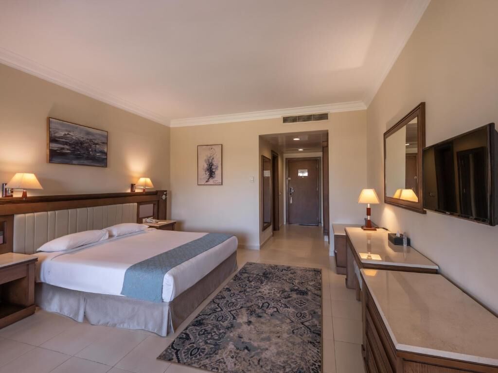 Une chambre d’hôtel avec un grand lit, des tables de chevet avec des lampes, un bureau avec une chaise, un miroir et une porte donnant sur une autre pièce.