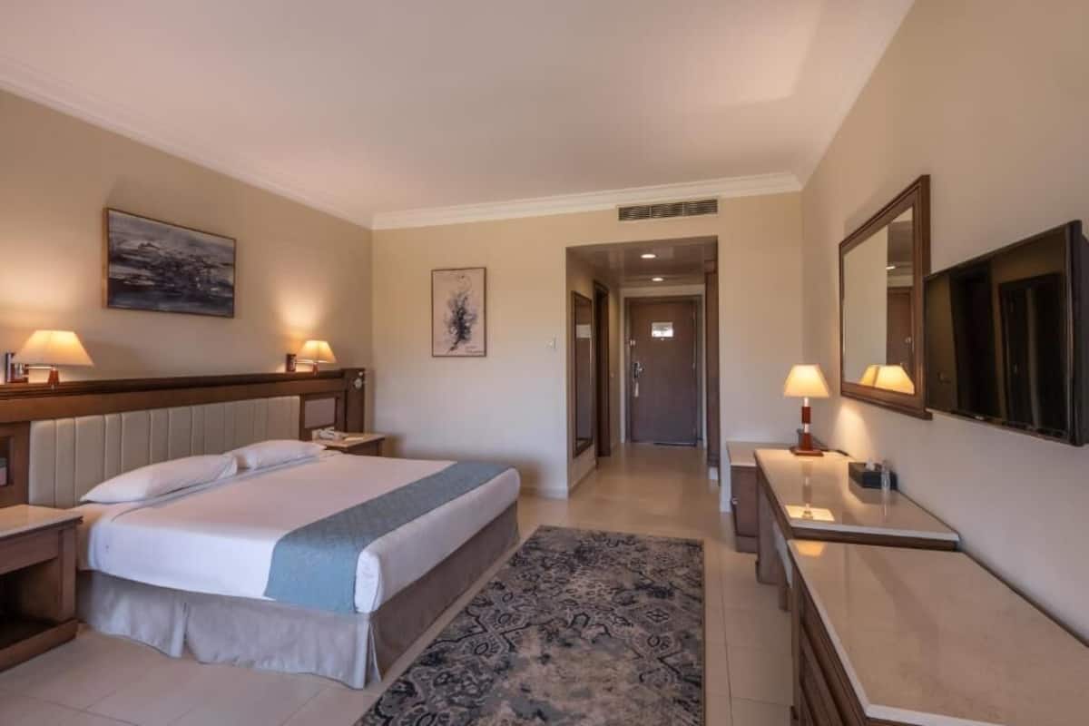 A hotel room with a bed, bedside tables, a desk, a mirror, and a door leading to another room.
