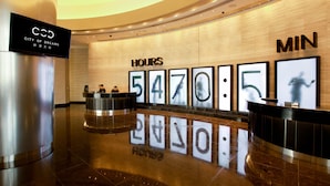 Zona de recepción de hotel con un reloj digital que muestra la hora, un mostrador de recepción y una pantalla grande sobre el mostrador.