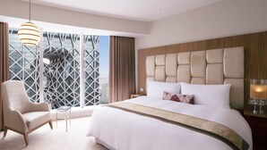Habitación de hotel moderna con una cama grande, una silla, una mesita y vistas a un paisaje urbano a través de amplios ventanales.