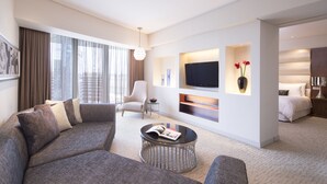 Una habitación de hotel moderna con un sofá grande, una mesita de centro redonda, un televisor de pantalla plana montado en la pared, una cama con una colcha blanca y un jarrón con flores sobre una mesita de noche.