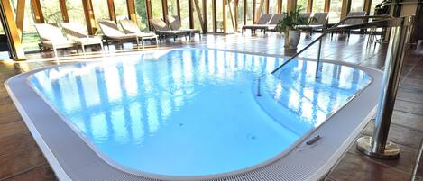 An indoor swimming pool with lounge chairs and a view of trees through large windows.