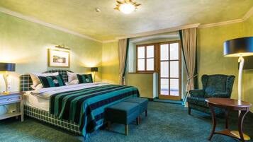 A hotel room with a bed, bedside table, lamp, chair, bench, and a window with curtains.
