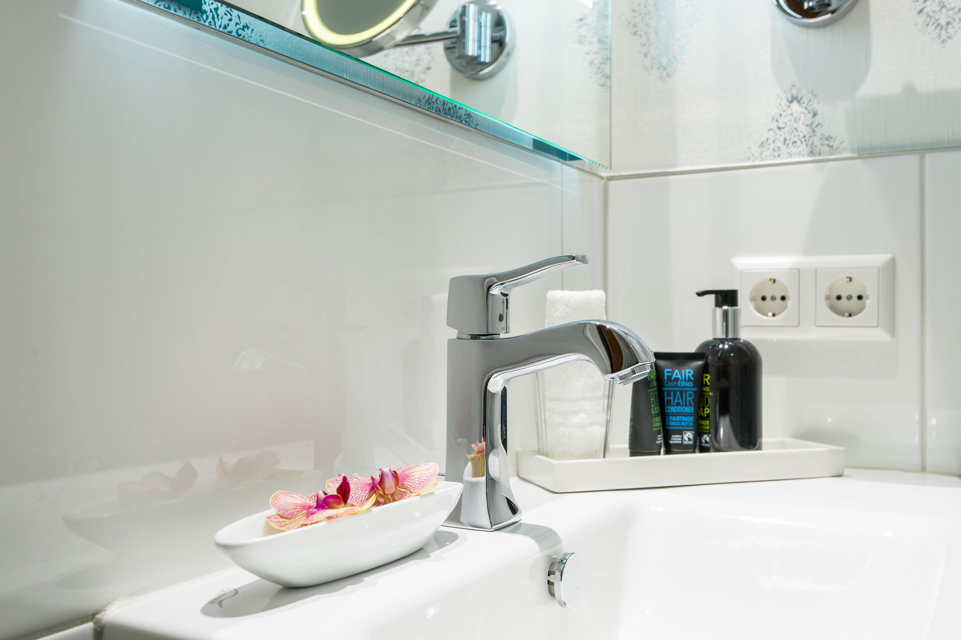 A modern bathroom with a white sink, a chrome faucet, and a glass bowl with flowers. There are two bottles of hair care products on the sink.