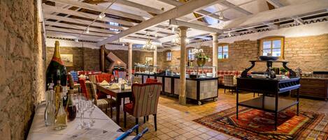 Un restaurante con vigas de madera a la vista, paredes de piedra y suelo de baldosas. Hay mesas con sillas, una zona de bar y una alfombra decorativa.