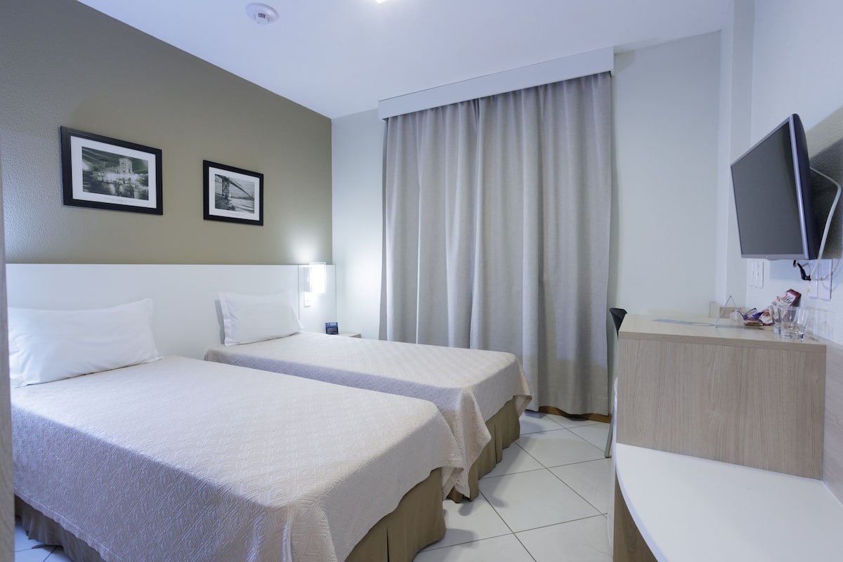 A hotel room with two beds, a desk, a TV, and a window with curtains.