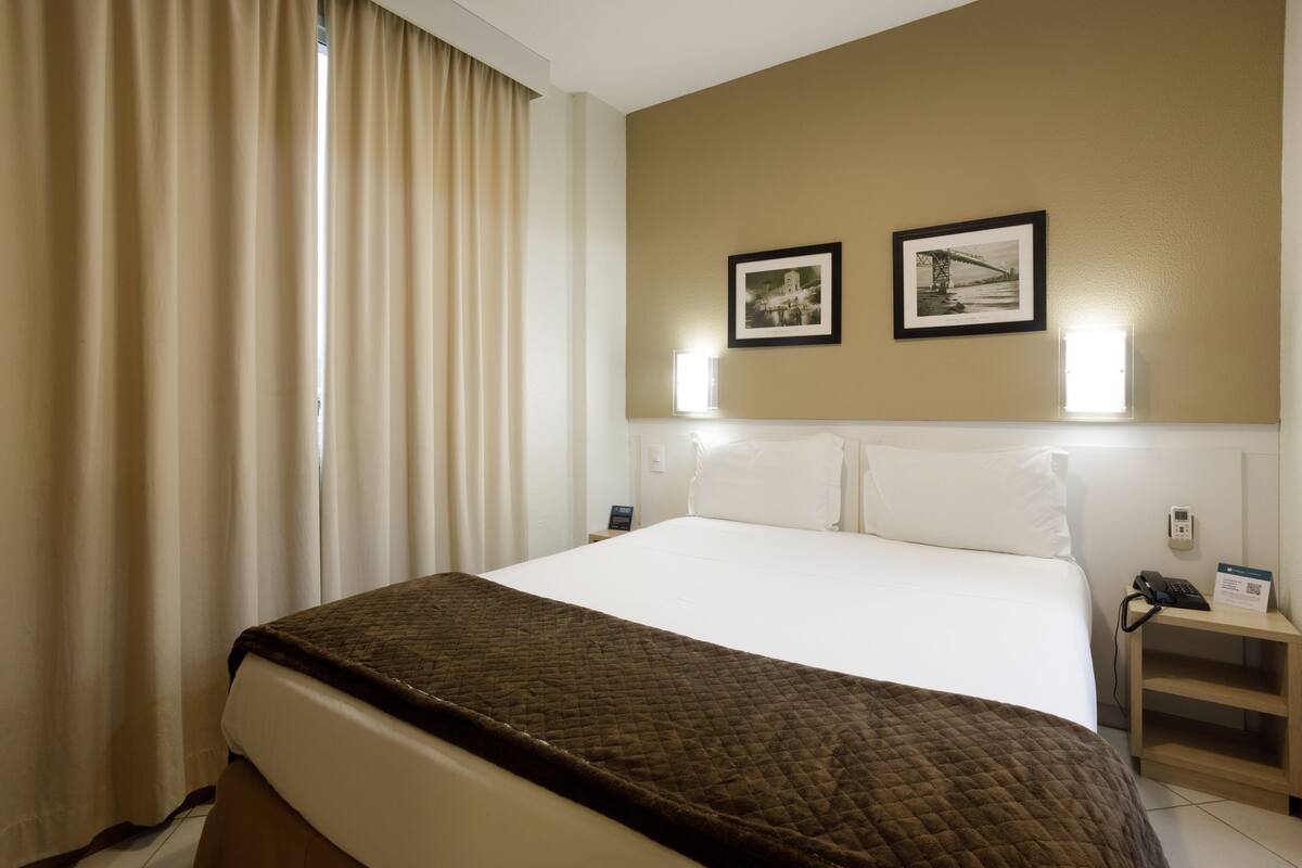A hotel room with a large bed, a bedside table with a phone and a lamp, and two framed pictures on the wall.
