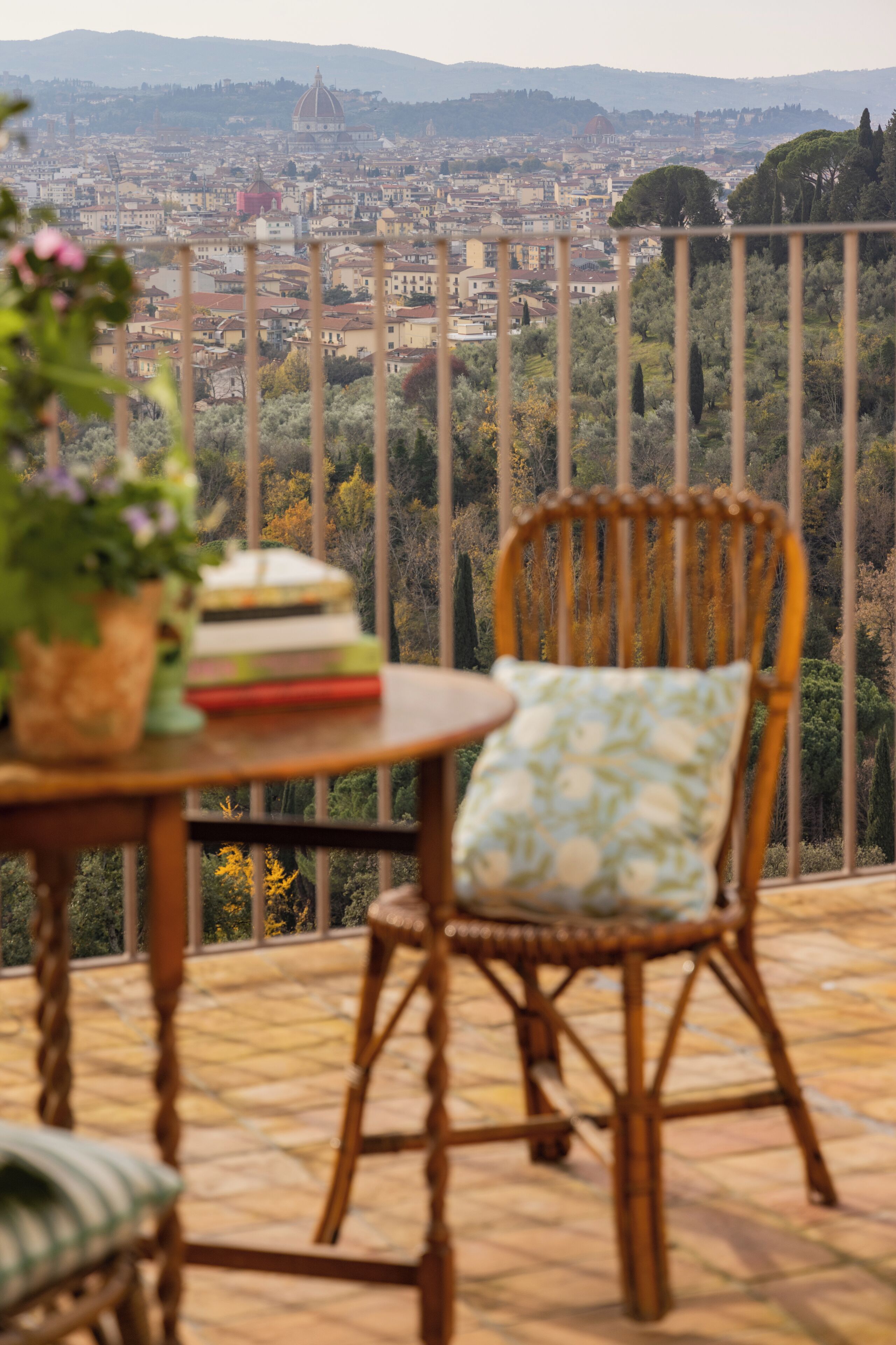 Una silla de mimbre con un cojín, una mesa con libros y una planta en maceta, y vista de un paisaje urbano con edificios y árboles.