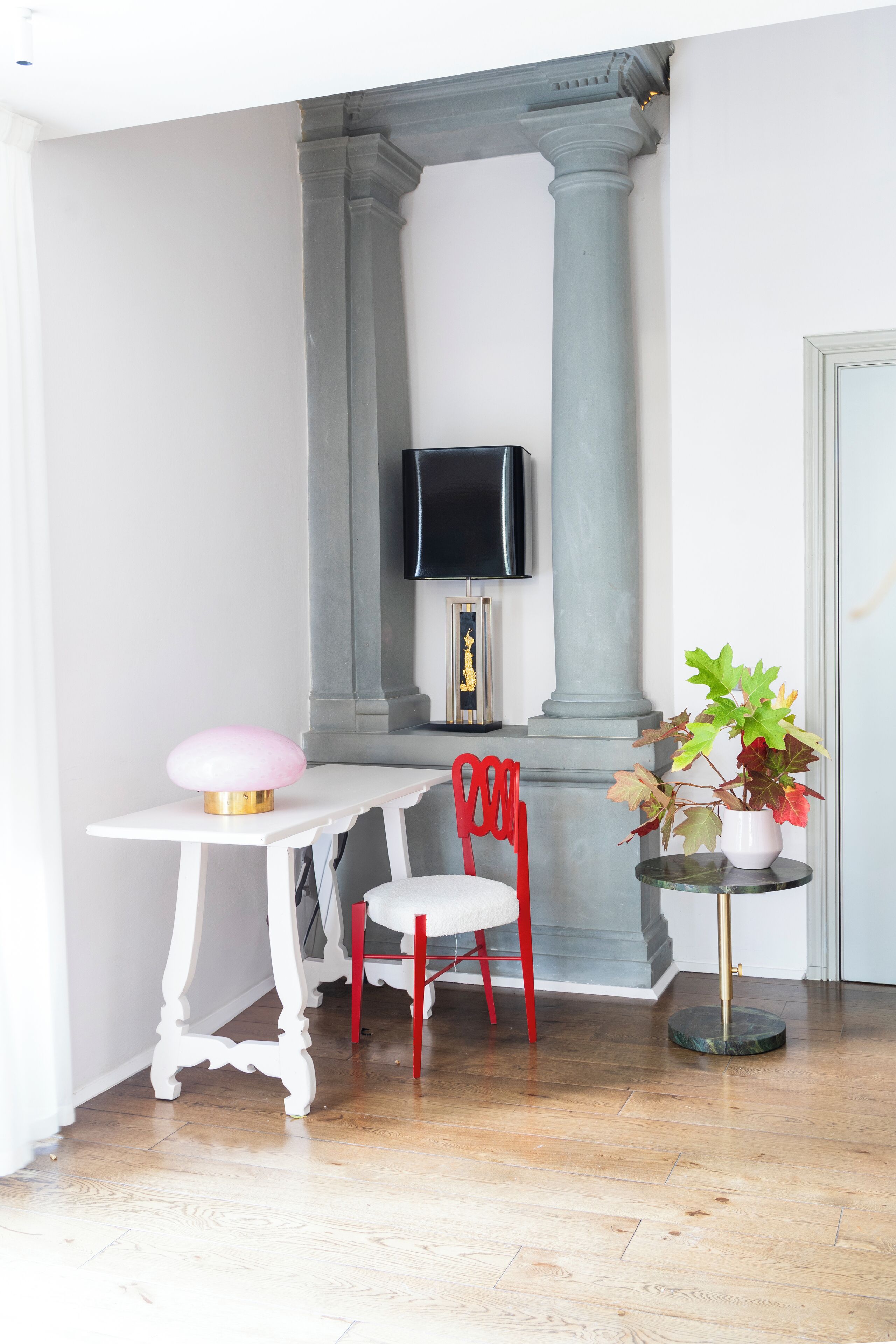 Una habitación con una mesa blanca, una silla roja, una lámpara rosa, una planta en maceta y un televisor montado sobre un pilar gris.