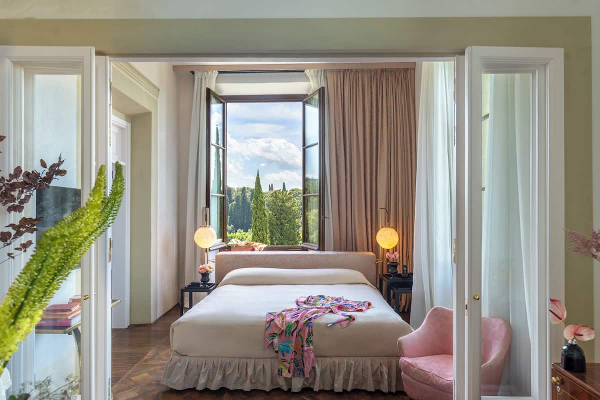 A spacious bedroom with a large bed, a view through an open window, and a pink chair.