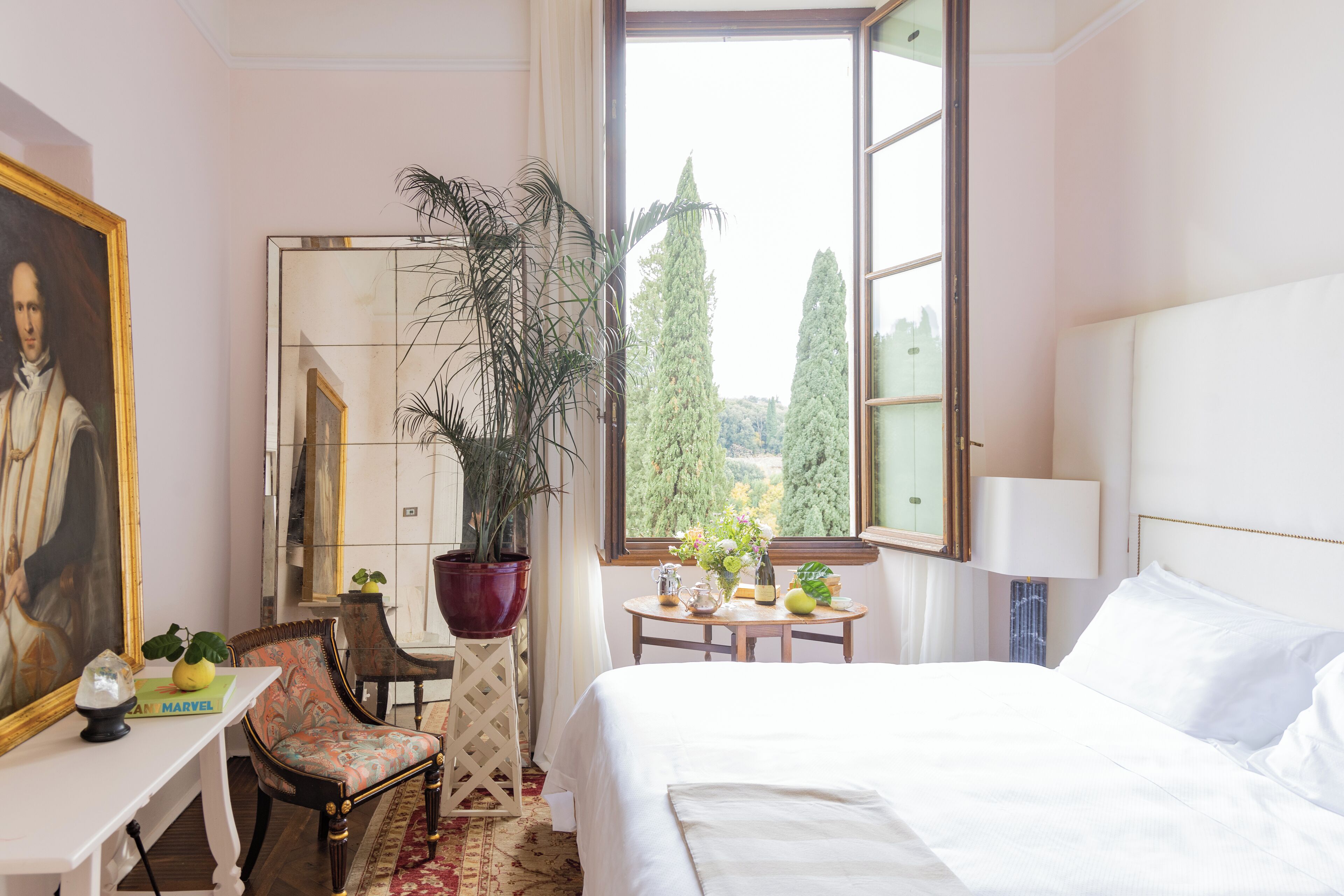 Hotelkamer met een bed, bureau, stoel, grote spiegel, een kamerplant en raam met uitzicht op bomen.