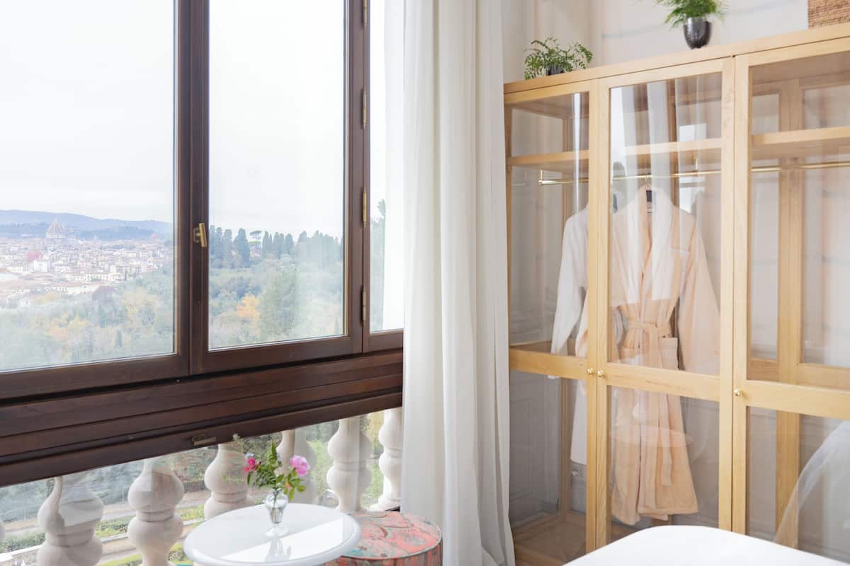 A room with a large window, a round table with a vase, and a wooden cabinet with a robe hanging inside.