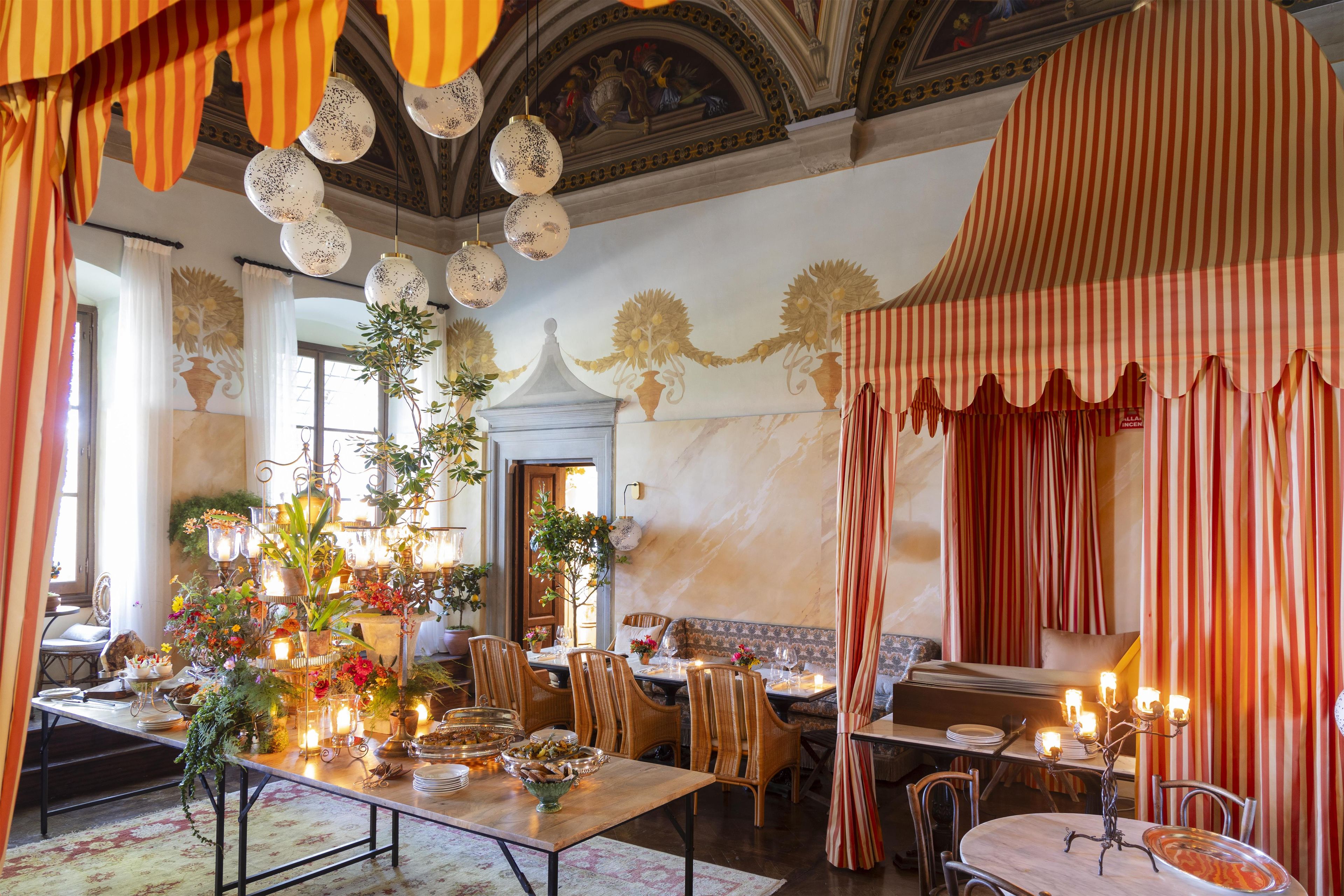Un comedor con una larga mesa dispuesta para una comida, rodeada de sillas, con un techo decorativo y un mural en la pared.