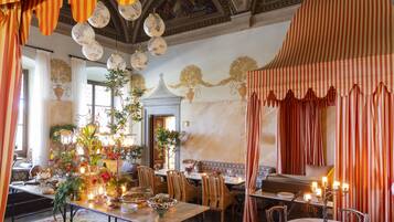 Un comedor con una larga mesa dispuesta para una comida, rodeada de sillas, con un techo decorativo y un mural en la pared.