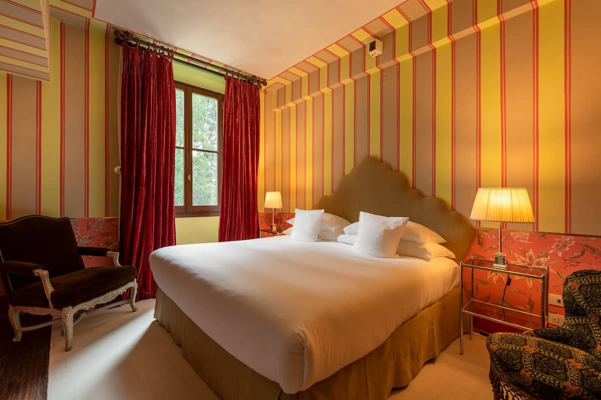 A hotel room with a large bed, two bedside lamps, a chair, and a window with red curtains.