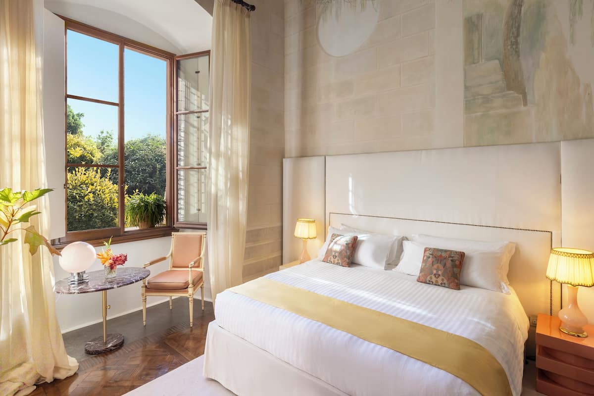 A hotel room with a large bed, a small table with a lamp, a chair, and a view of greenery outside.