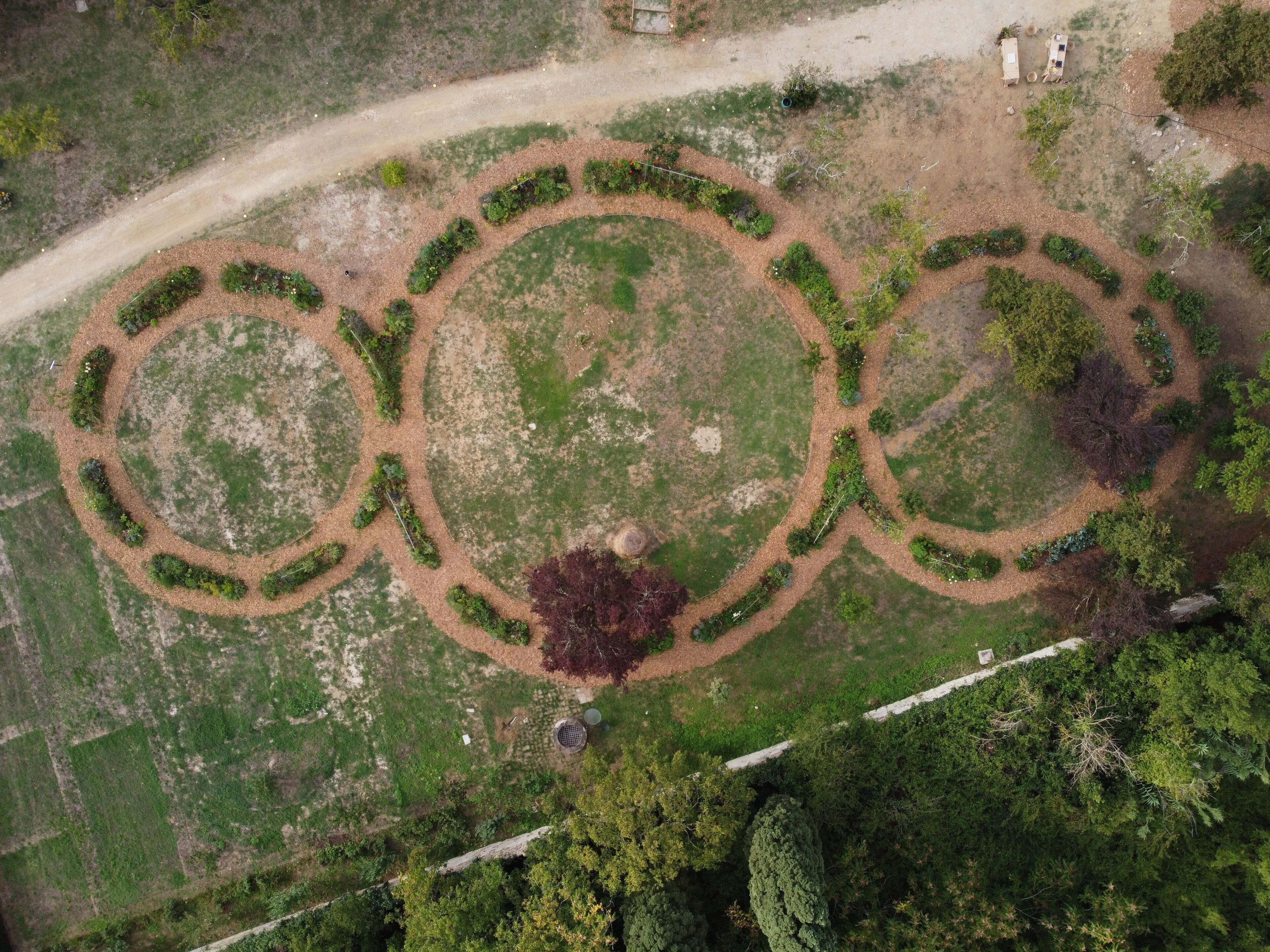 An aerial view of a landscaped area with three circular garden beds, a dirt path, and a small building.