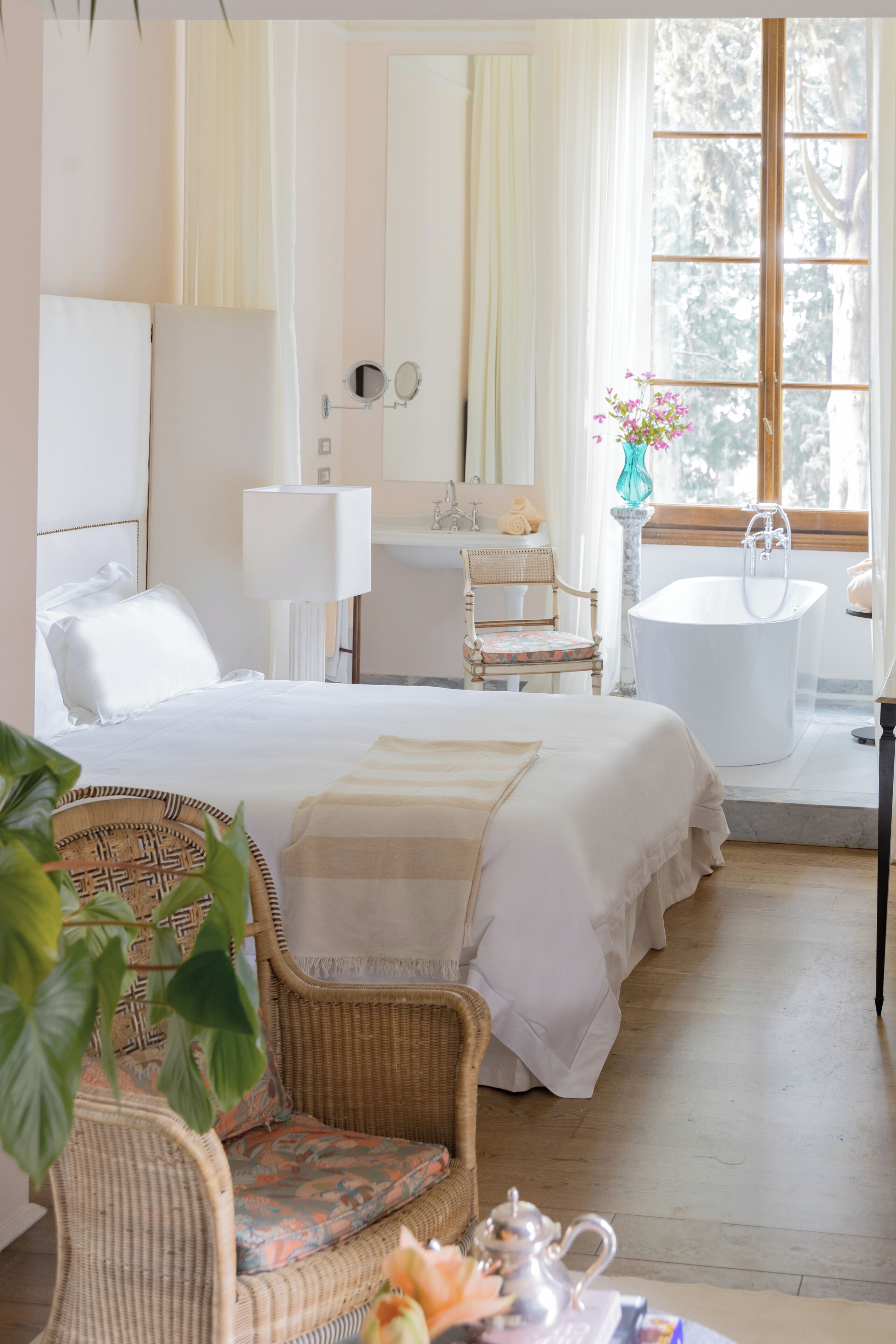 Una habitación de hotel con una cama, una silla, una mesa con tetera, una bañera y un ventanal con cortinas.