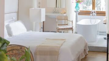 Una habitación de hotel con una cama, una silla, una mesa con tetera, una bañera y un ventanal con cortinas.