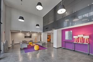 Een ruime lobby met een hoge plafond, moderne hanglampen, een accentmuur in paars, en een zithoek met een paars tapijt.