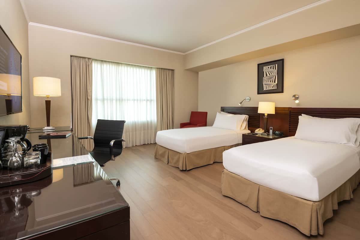 A hotel room with two beds, a desk with a lamp, a chair, and a window with curtains.