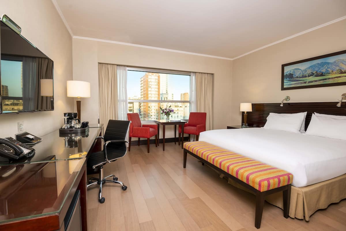 A hotel room with a large bed, a desk with a telephone, a chair, a table with a vase, and a window with a city view.