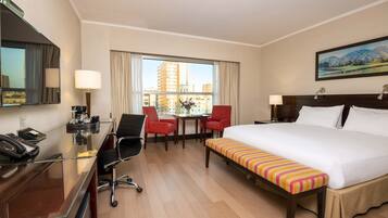 A hotel room with a large bed, a desk with a telephone, a chair, a table with a vase, and a window with a city view.