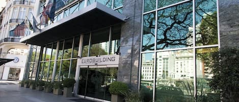 A modern building with a large glass facade, a sign reading 'EUROBUILDING,' and a reflection of trees in the windows.