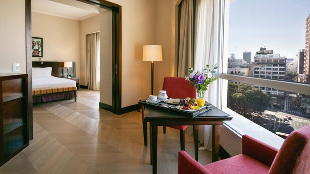 Habitación de hotel con cama, mesa de desayuno, una silla, lámpara y vista de la ciudad.