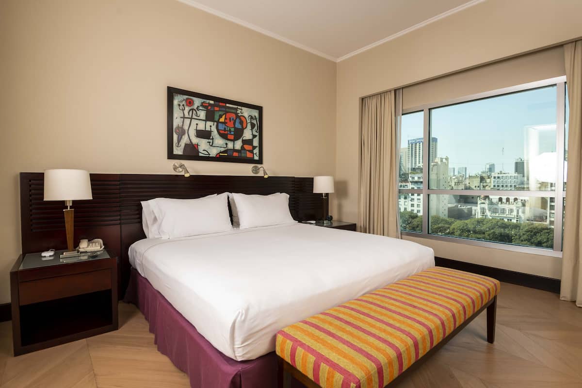 A hotel room with a large bed, two bedside tables, a bench, a city view, and a painting on the wall.