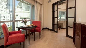 Habitación de hotel con comedor, una silla roja, una mesa con un jarrón de flores y vista a un edificio a través de la ventana.
