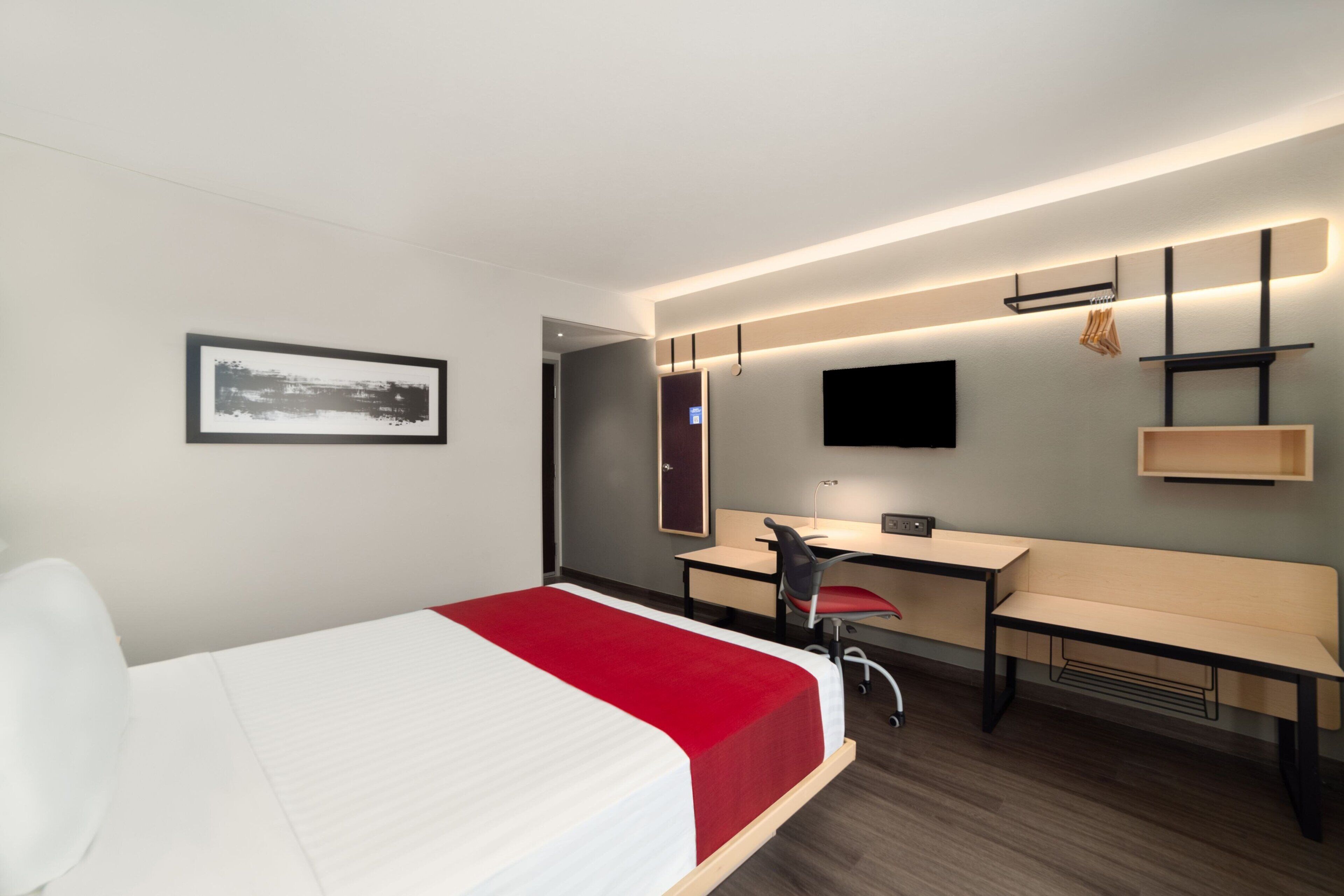 A modern hotel room with a bed, desk, chair, and a TV mounted on the wall.