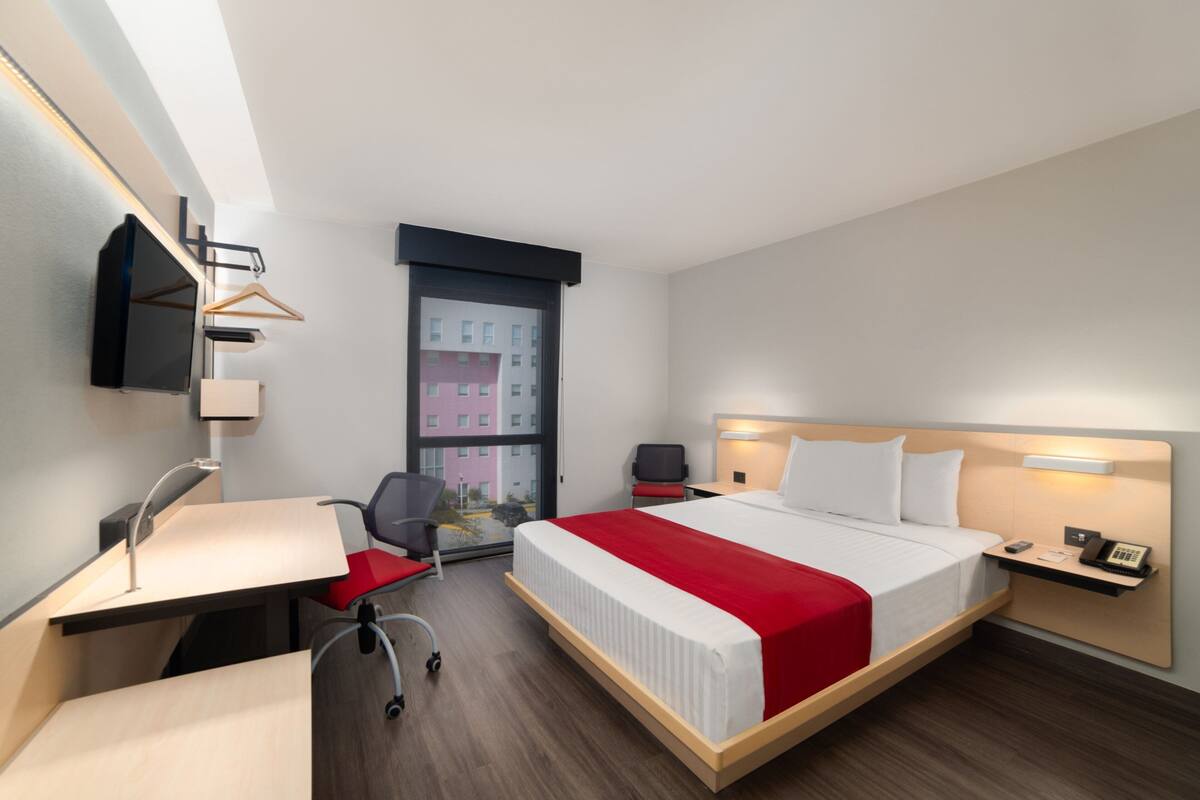 Habitación de hotel moderna con una cama grande, un escritorio con silla, una mesita y un ventanal con vistas a edificios.