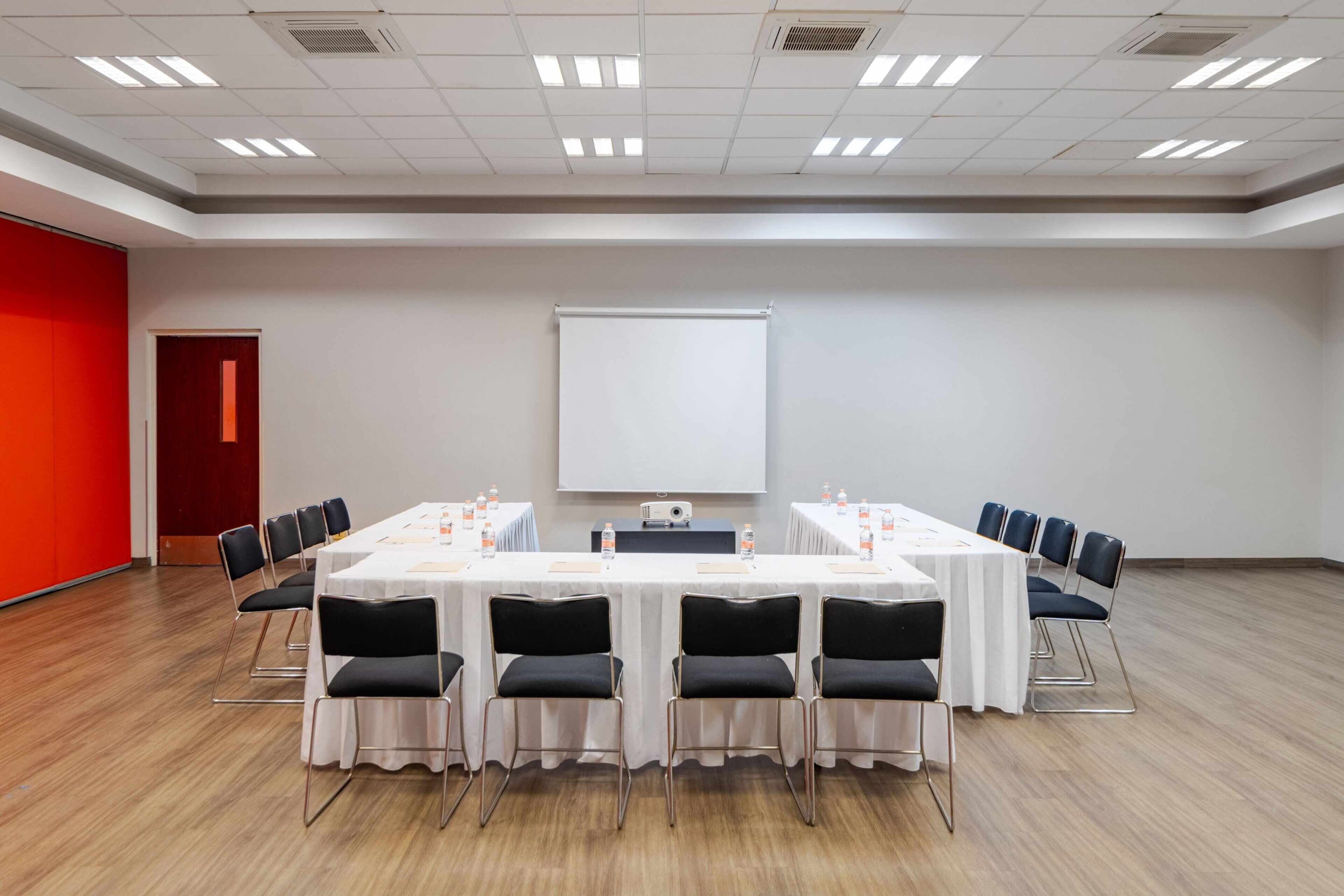 Un salón de conferencias con una mesa blanca, sillas negras, una pantalla de proyector y una pared roja.