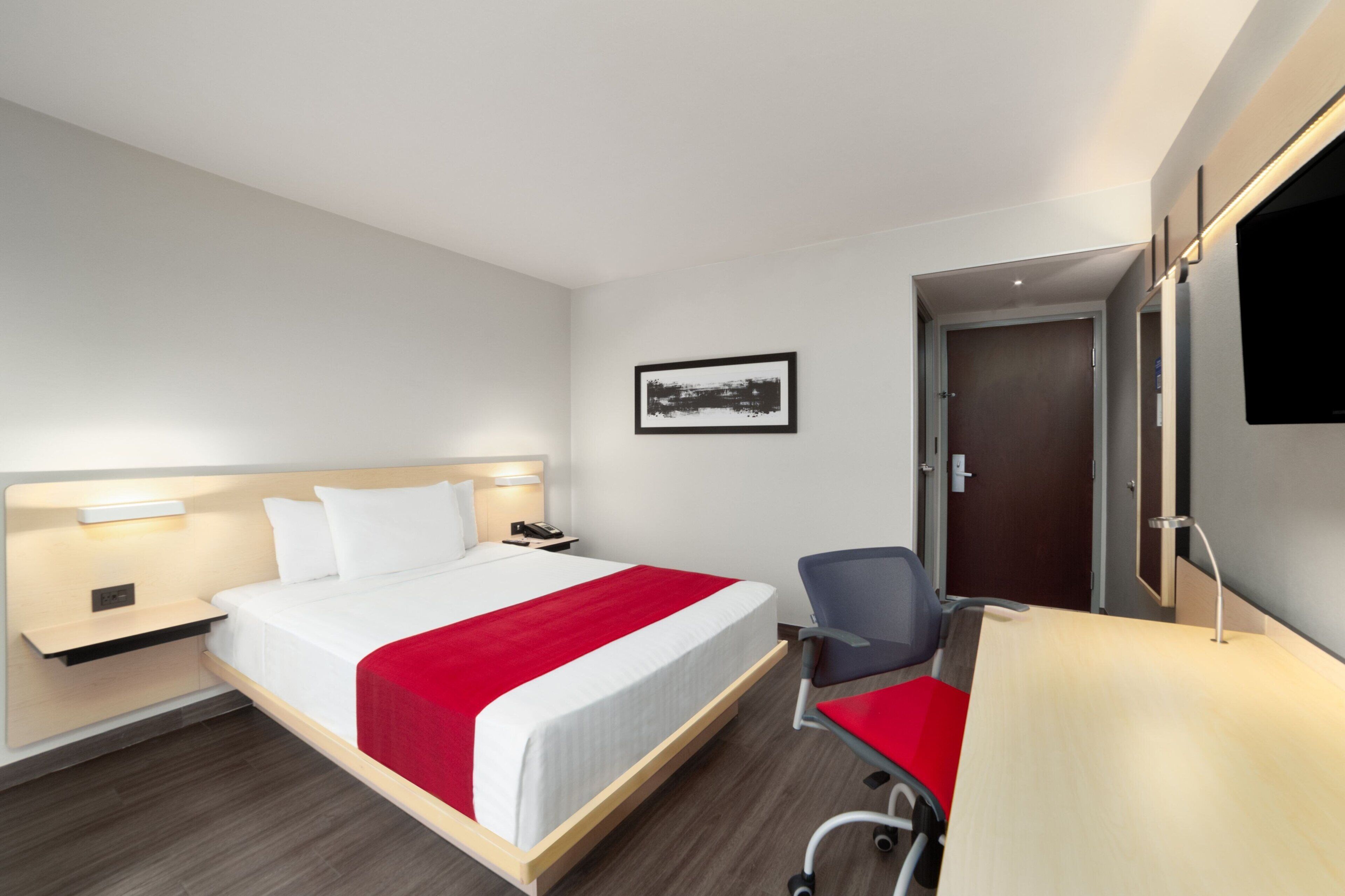 A hotel room with a bed, a desk, a chair, a TV, and a door.