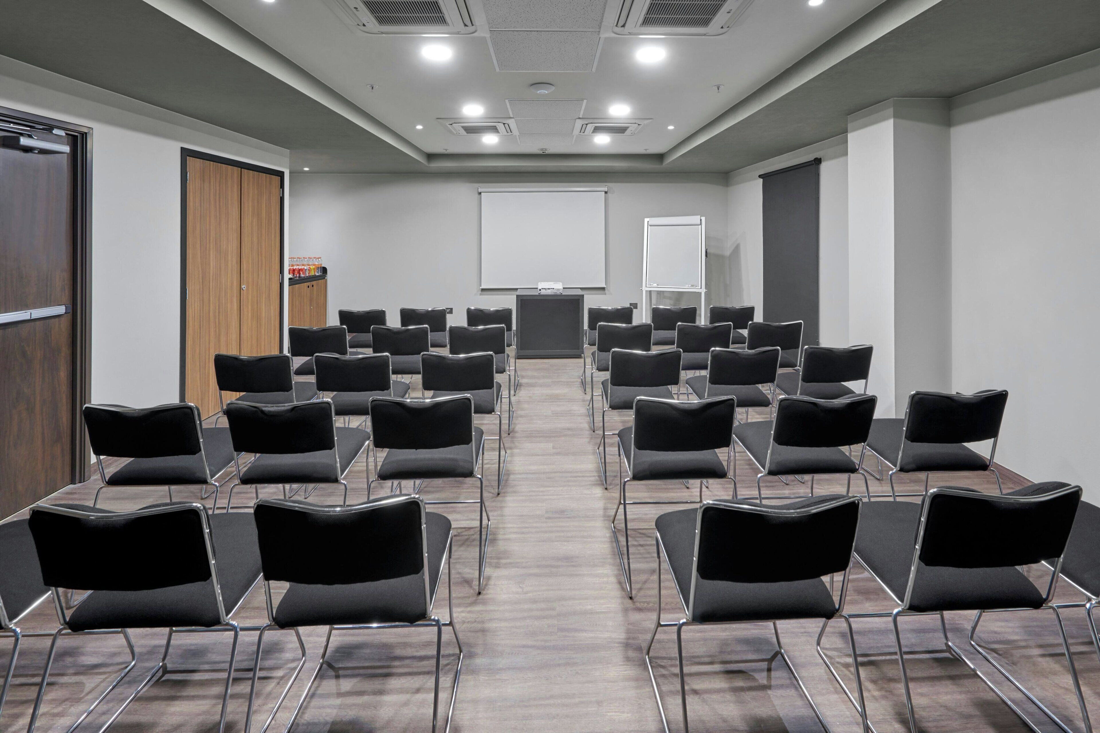 Un salón de conferencias con hileras de sillas negras, una pantalla de proyector y una pizarra.