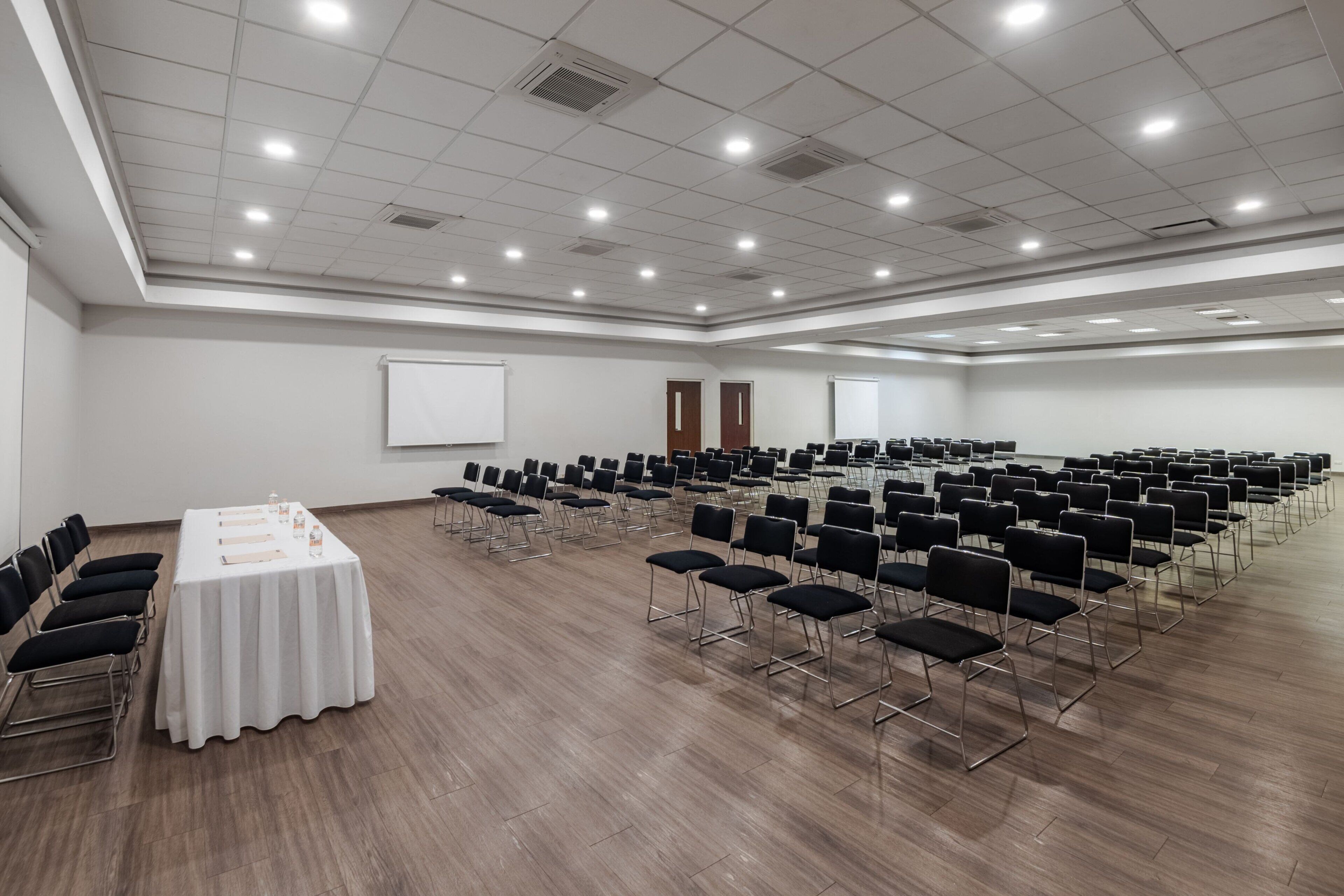 Une grande salle de conférence avec des rangées de chaises, une longue table recouverte d’une nappe blanche et un écran de projection.