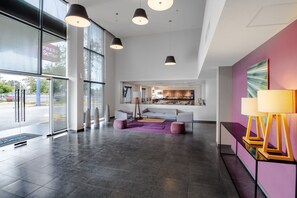 Un vestíbulo amplio con diseño moderno, que cuenta con una zona de estar grande con un sofá y sillones, una mesa de centro y una alfombra colorida. Hay lámparas colgantes del techo y un ventanal que proporciona luz natural.
