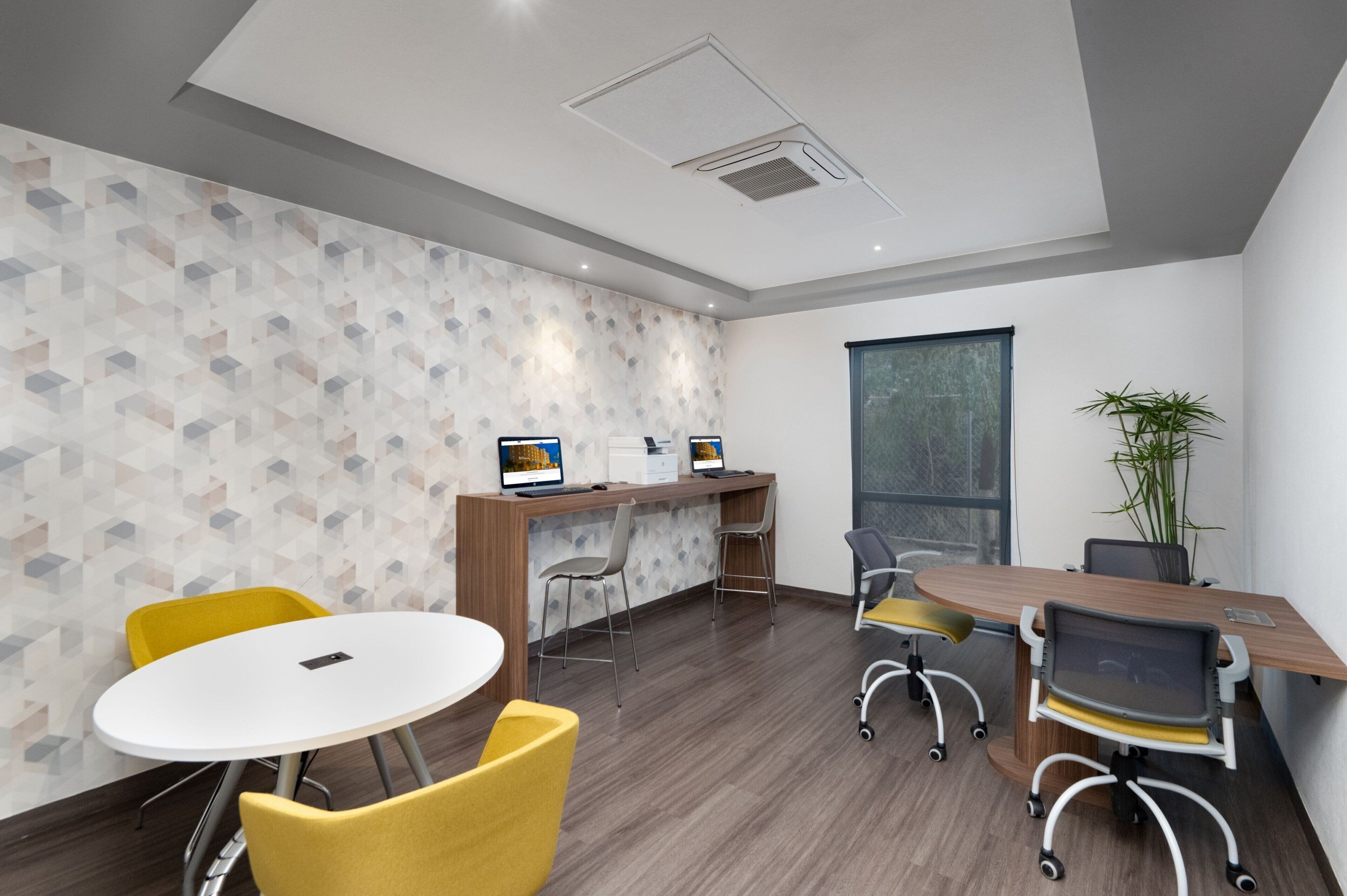 A modern meeting room with a wooden desk, two computers, yellow chairs, and a large window.