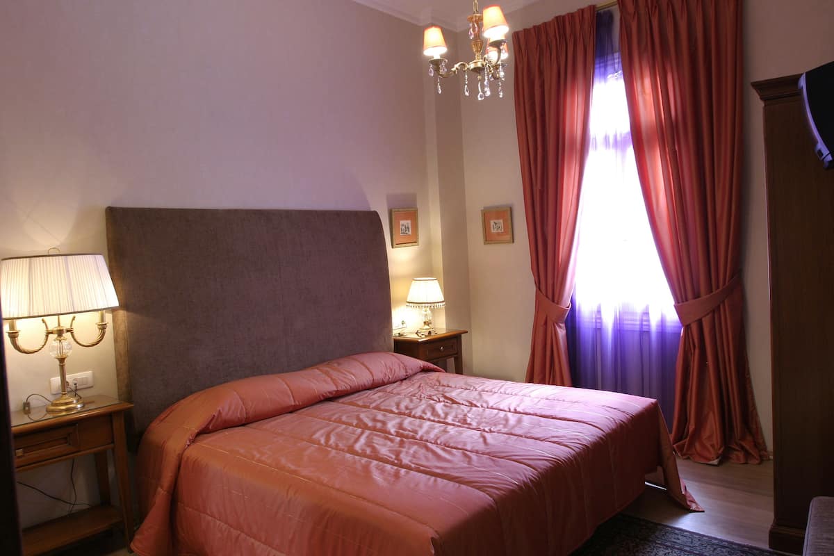 Ein Schlafzimmer mit einem großen Bett, einem Nachttisch mit Lampe, einer Kommode und einem Fenster mit Vorhängen.