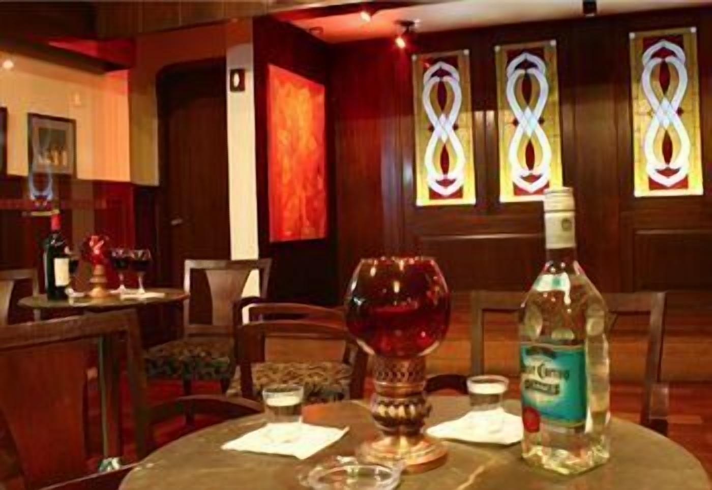A hotel room with a round table, a bottle of Cointreau, a glass of red wine, and a candle. There are three stained glass windows with a repeating pattern.