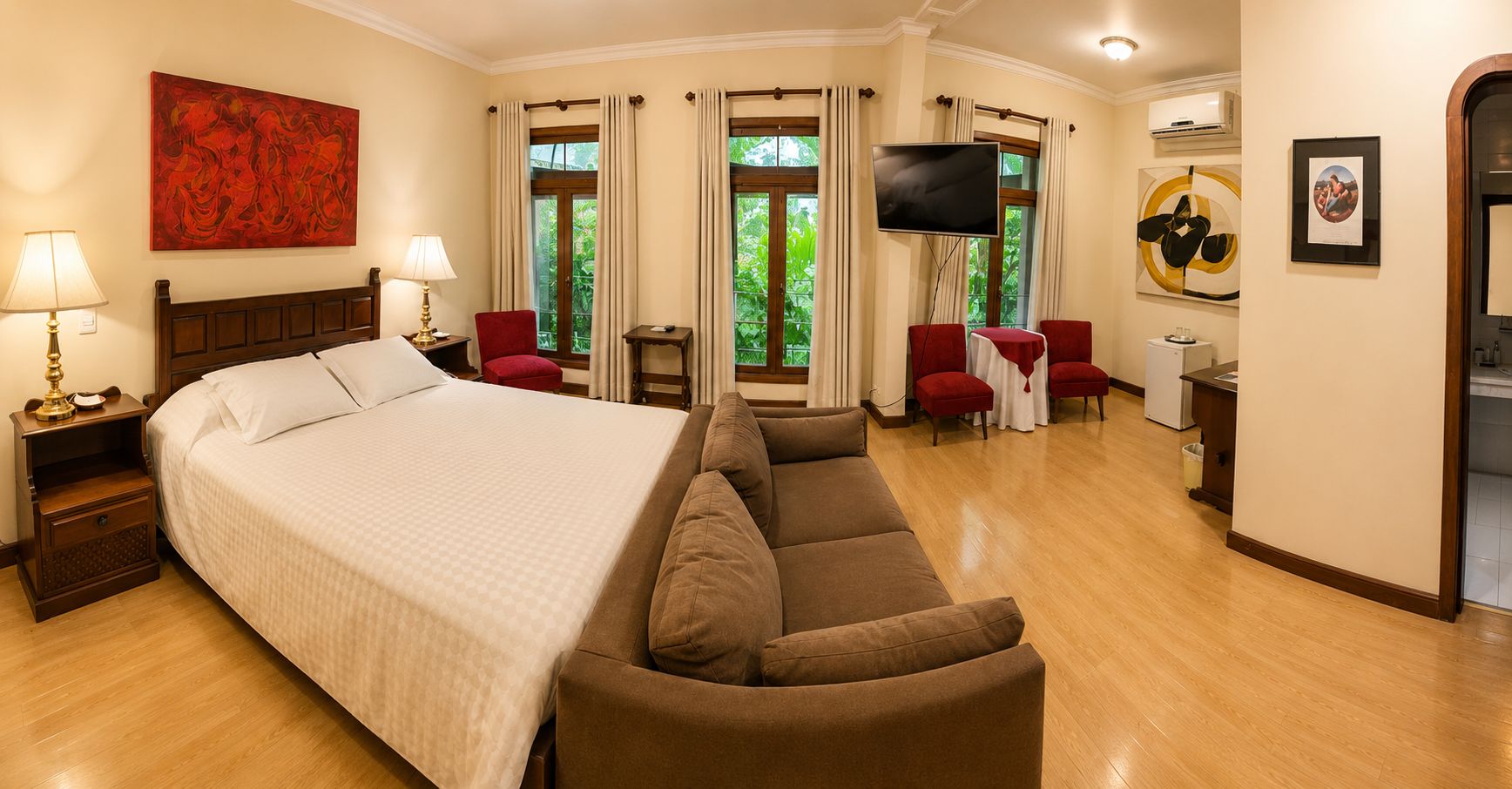 A hotel room with a large bed, a sofa, a TV, and a dining area with chairs.