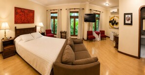 A hotel room with a large bed, a sofa, a TV, and a dining area with chairs.
