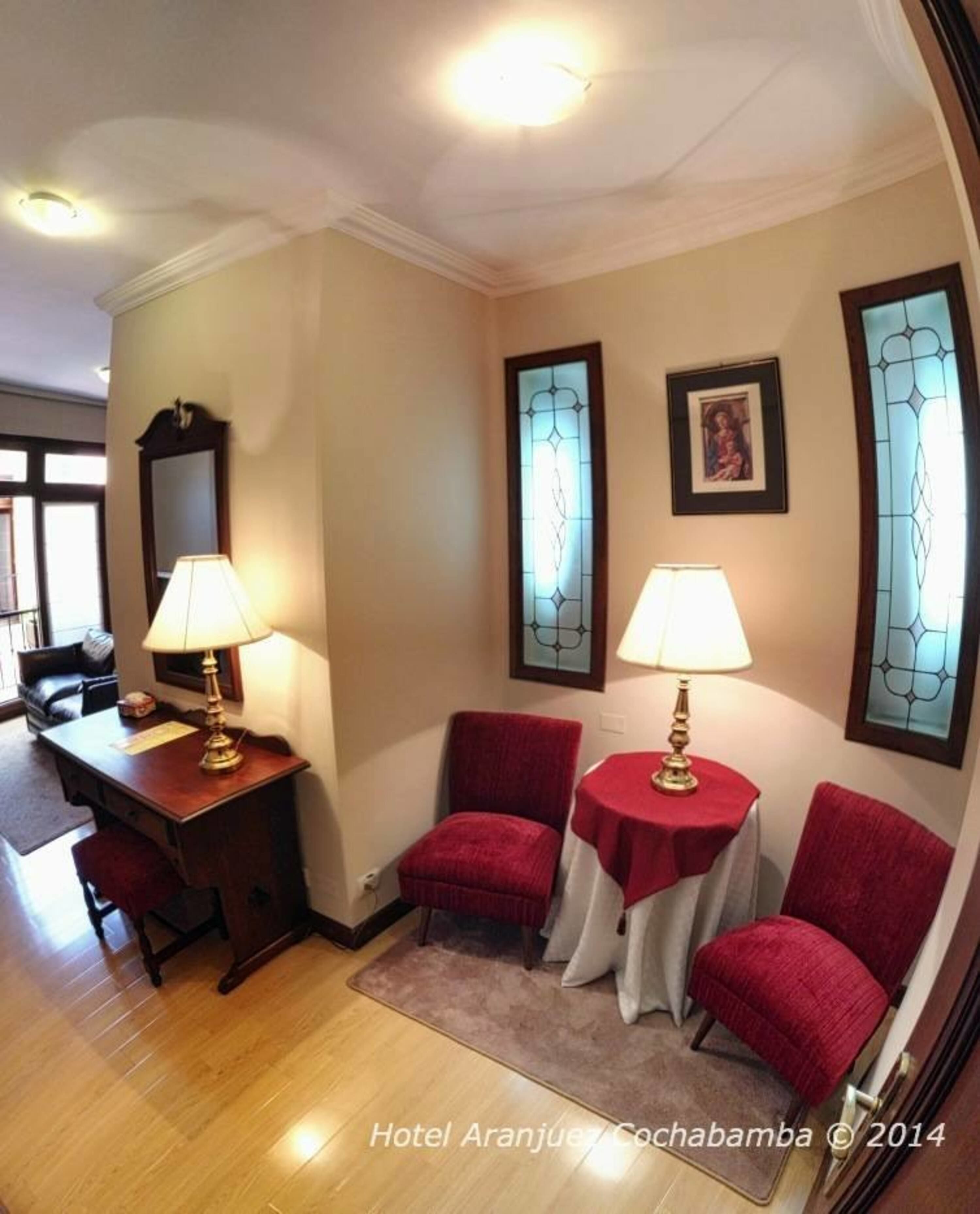 A hotel room with a desk, two red chairs, a small table with a lamp, and a stained glass window.