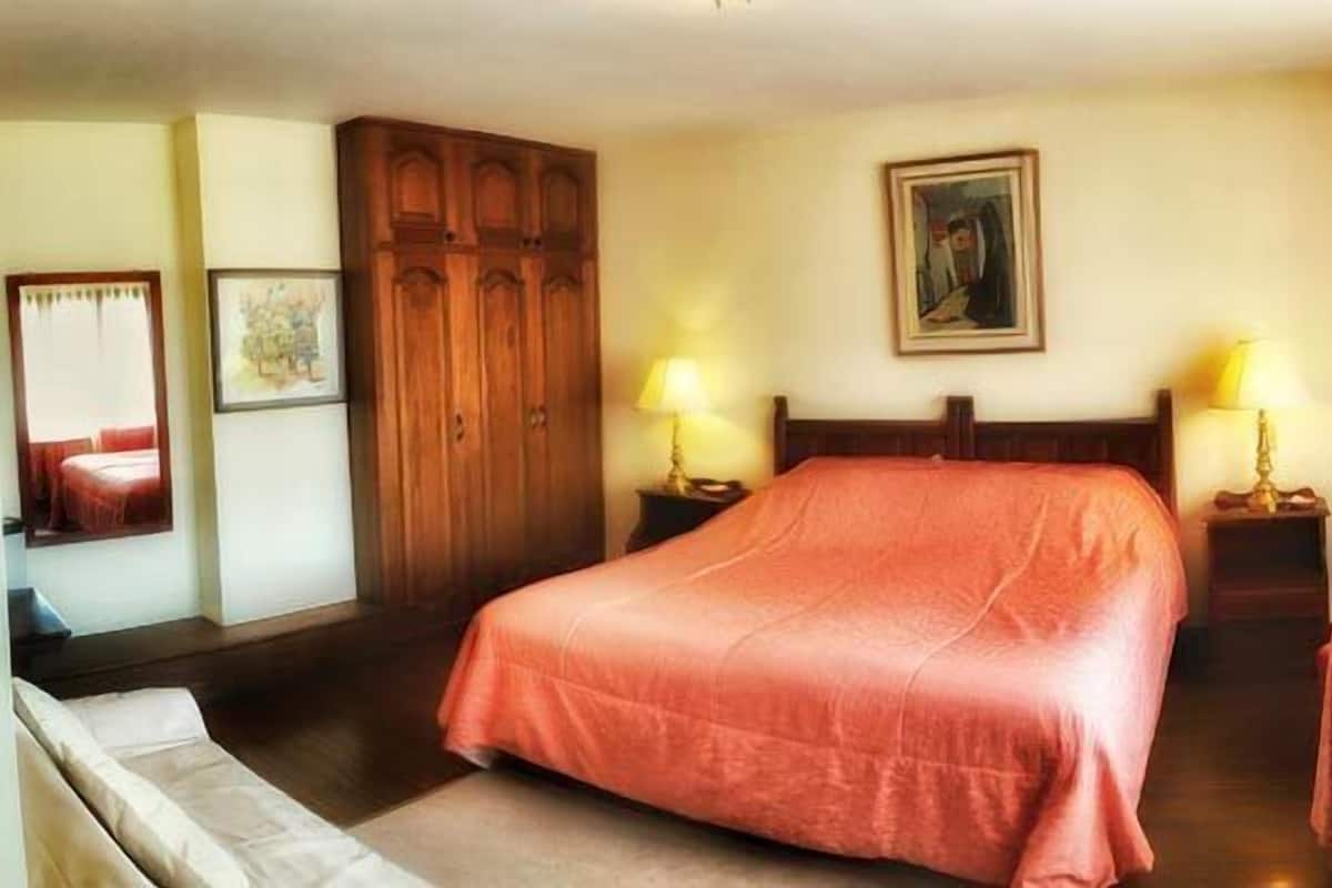 Una habitación de hotel con una cama grande, dos mesitas de noche con lámparas, una mesa redonda, una silla, un armario y un cuadro en la pared.