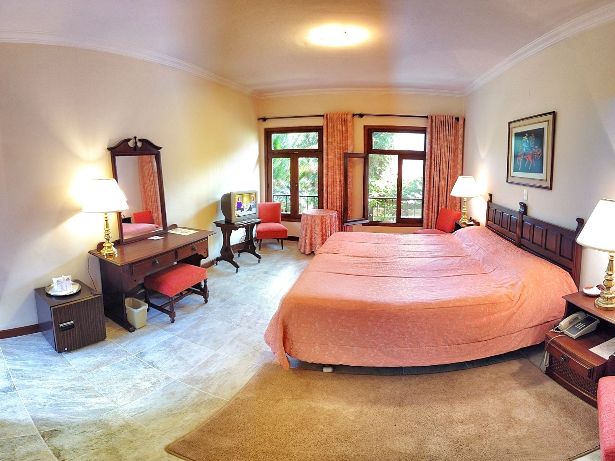 Habitación de hotel con una cama grande, un escritorio con silla, una mesita, un espejo y una ventana con cortinas.