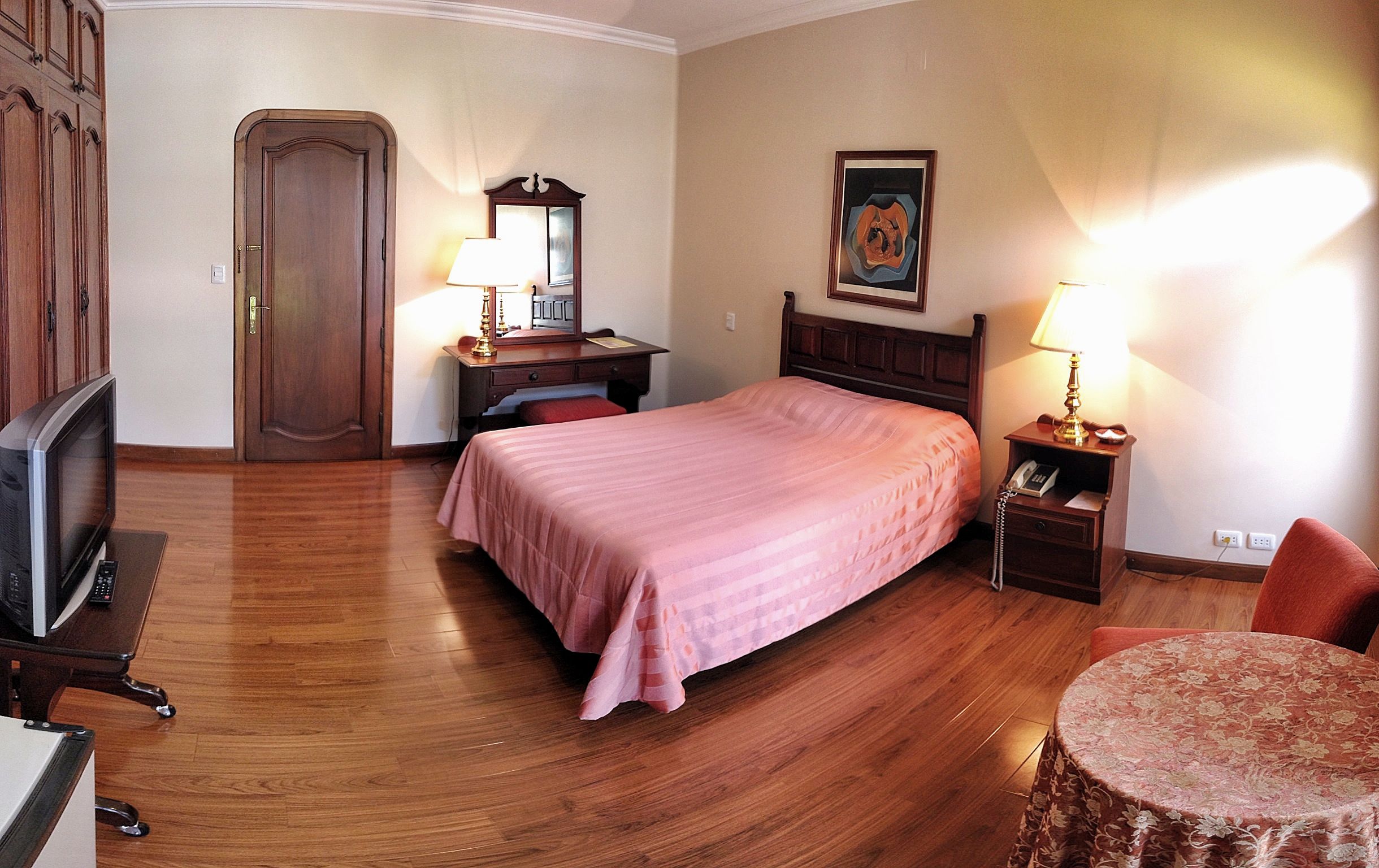 A hotel room with a bed, a desk with a lamp, a television, and a wooden wardrobe.