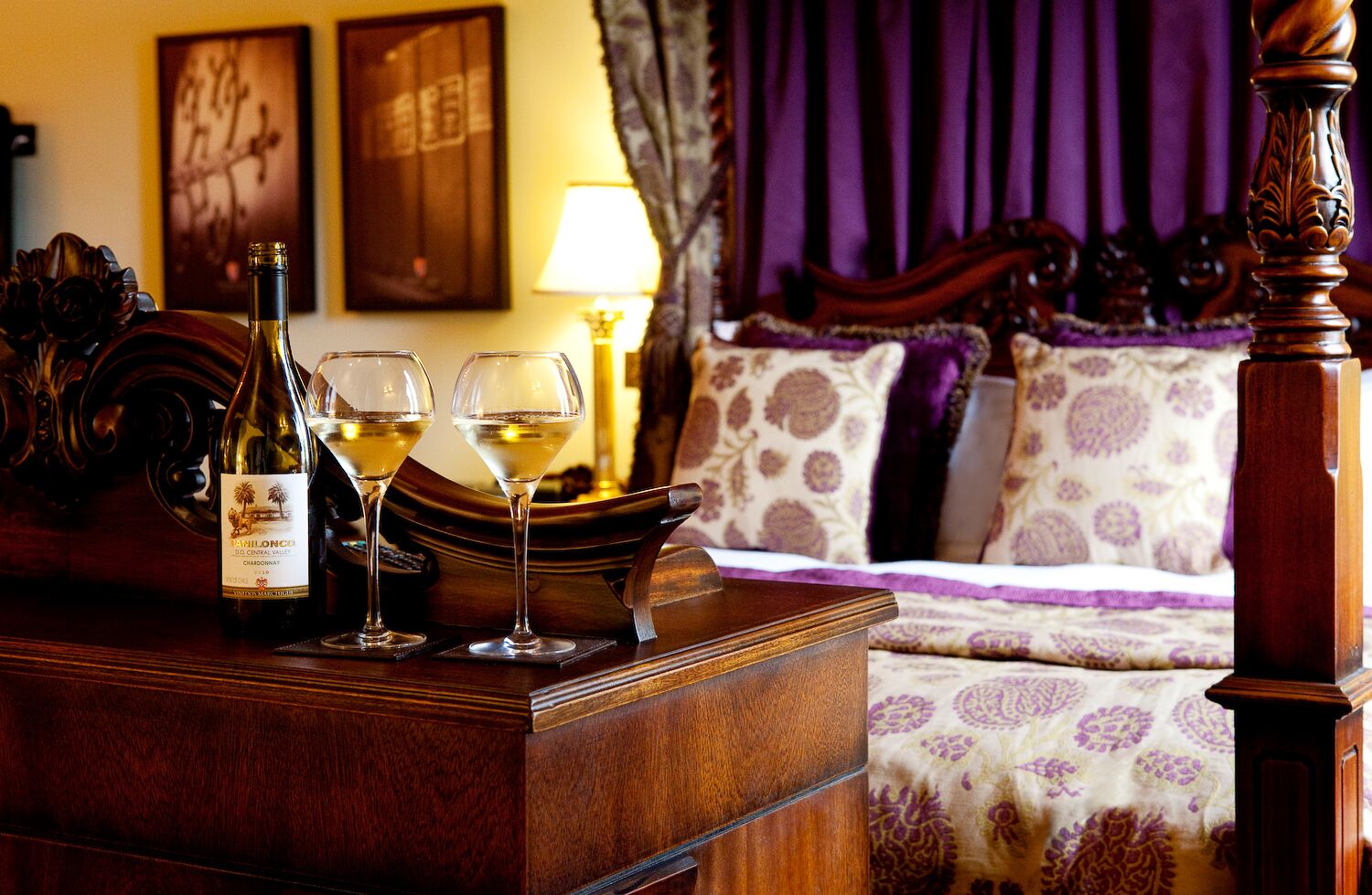 A wooden table with a bottle of wine and two glasses, a bed with patterned bedding, and a lamp in a room with purple curtains.