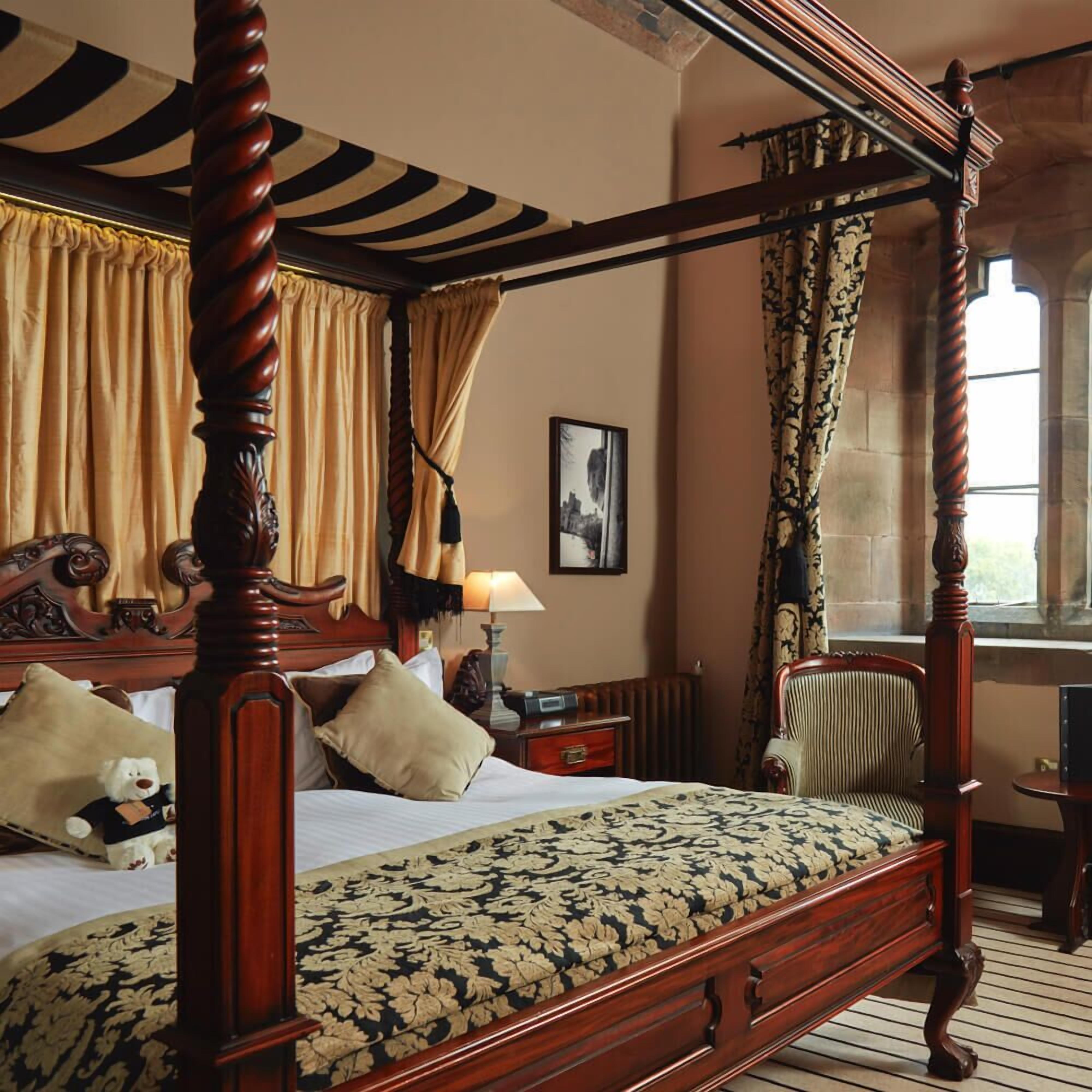 A four-poster bed with a floral bedspread, a wooden nightstand with a lamp, a chair, and a framed picture on the wall.