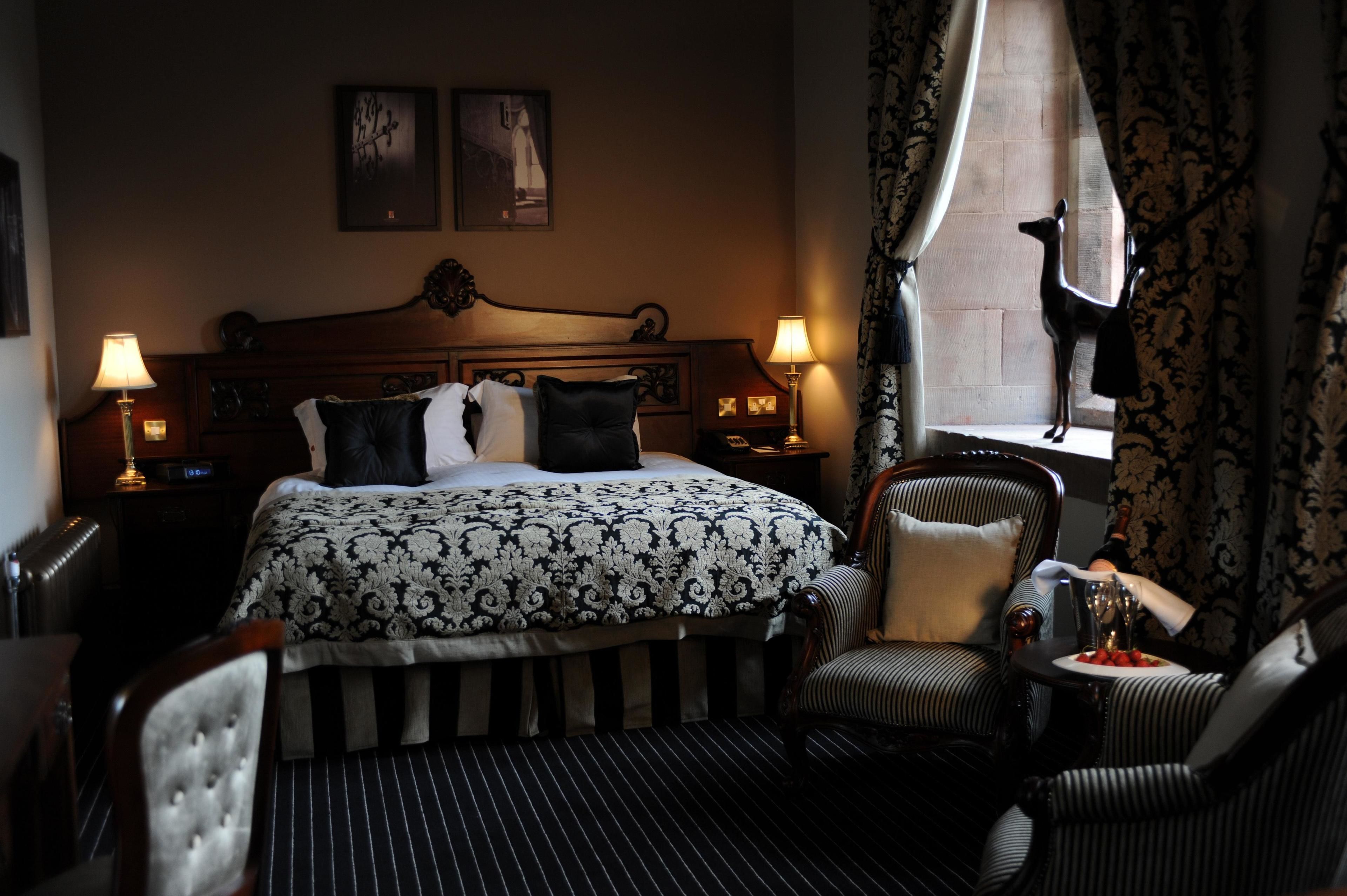 A hotel room with a large bed, two bedside lamps, a chair, a small table with a plate of strawberries, and a window with curtains.